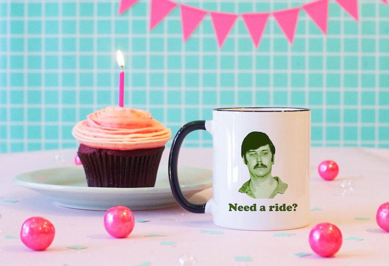 May include: White coffee mug with a black handle and a black rim. The mug has a green image of a man with a mustache and glasses and the text "Need a ride?" printed on it. The mug is sitting on a pink table with pink and white confetti.