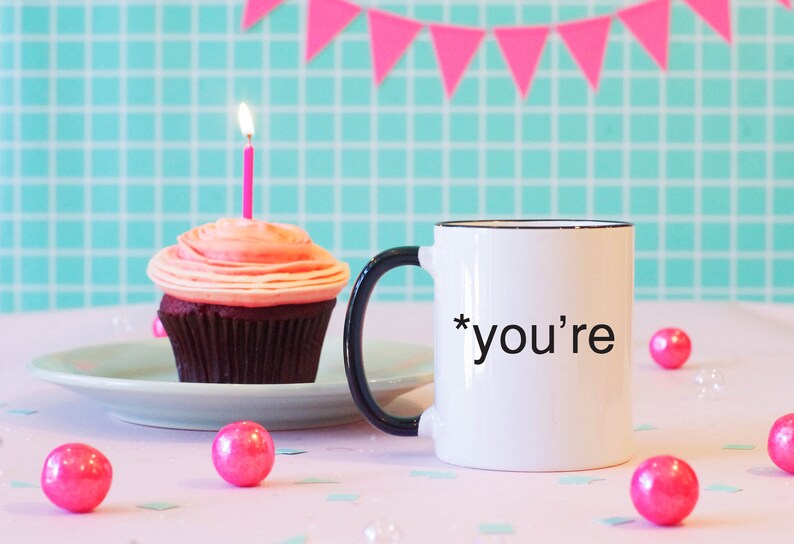 May include: White coffee mug with a black handle and black rim. The mug has the text "*you're" printed on it. A pink cupcake with a lit candle is on a plate in the background.