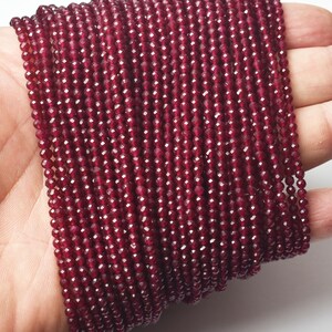 May include: A collection of deep red, faceted, round beads strung together in multiple parallel strands. Each small bead features a reflective, multi-faceted surface that catches the light, creating a uniform, dark crimson appearance across the entire group.