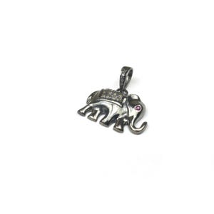 May include: A silver-toned elephant pendant with a small red gem for an eye. The elephant has a decorative blanket and is adorned with small, clear stones. The pendant is on a white background.