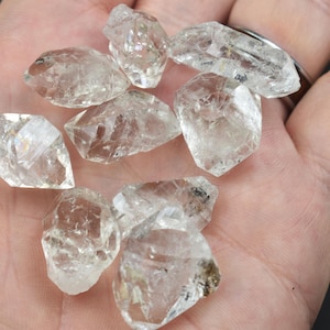 Puede incluir: Una colección de pequeños cristales de cuarzo en bruto, transparentes, con formas irregulares y facetadas y bordes afilados. Estas piedras translúcidas presentan fracturas internas naturales y diversos grados de claridad, reflejando la luz en sus superficies cristalinas irregulares.