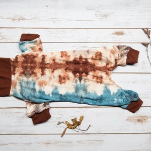 May include: A brown and blue tie-dye dog jumpsuit with brown trim. The jumpsuit is laid out flat on a white wooden surface.