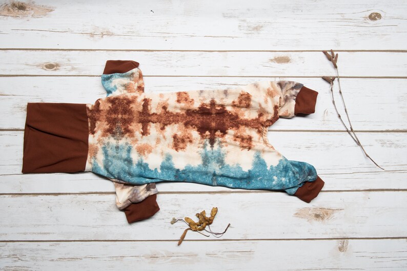 May include: A brown and blue tie-dye dog jumpsuit with brown trim. The jumpsuit is laid out flat on a white wooden surface.