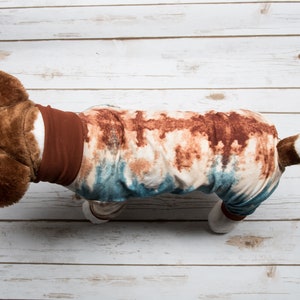 May include: A brown and white dog wearing a blue and brown tie-dye pajama onesie with a brown collar.