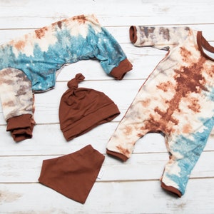 May include: A set of baby clothes in a tie-dye pattern with brown accents. The set includes a long-sleeved jumpsuit, a hat, and a bib.