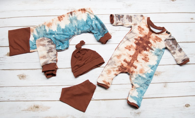 May include: A set of baby clothes in a tie-dye pattern with brown accents. The set includes a long-sleeved jumpsuit, a hat, and a bib.