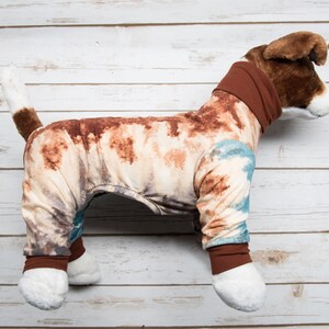 May include: A brown and white dog wearing a brown, blue, and white tie-dye pajamas. The pajamas have brown cuffs and a brown turtleneck.