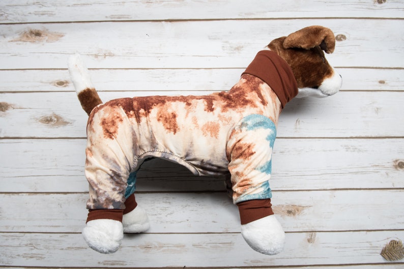 May include: A brown and white dog wearing a brown, blue, and white tie-dye pajamas. The pajamas have brown cuffs and a brown turtleneck.