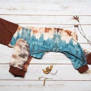 May include: A brown and blue tie-dye dog jumpsuit with brown cuffs. The jumpsuit is laid out flat on a white wooden surface.