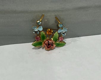 May include: A floral brooch featuring a central pink rose surrounded by green leaves and light blue flowers. The brooch has gold-colored accents and is adorned with small, colorful gemstones.