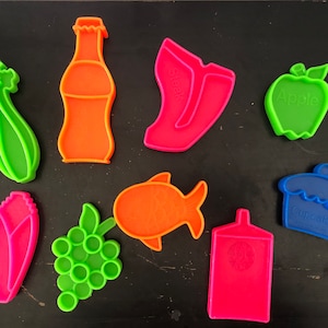 May include: A set of nine colorful plastic cookie cutters shaped like a bottle of soda, celery, a shoe, an apple, a fish, grapes, a corn cob, a cupcake, and a container.