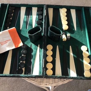 Puede incluir: Juego de backgammon abierto con una superficie de juego verde, puntas negras y crema, y ​​piezas de juego negras y crema. Incluye dados, cubiletes y un folleto con la etiqueta "BACKGAMMON RULES". El juego está abierto sobre una mesa de mimbre.