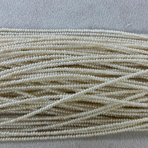 Peut inclure: Gros plan sur une collection de petites perles rondes blanc cassé. Les perles sont enfilées ensemble, créant une surface dense et texturée. Les brins sont superposés, suggérant une grande quantité de perles.