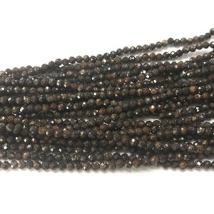 Puede incluir: Una hebra de cuentas de piedras preciosas facetadas marrones. Las cuentas son pequeñas y tienen una superficie rugosa y texturizada.