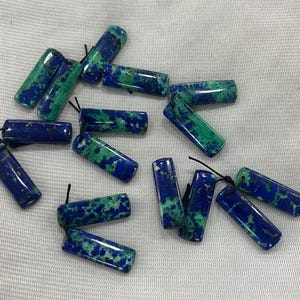 Può includere: Una collezione di 14 ciondoli in pietre preziose verdi e blu con anelli di corda neri. I ciondoli sono rettangolari e hanno un motivo maculato.