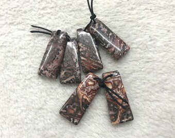 Puede incluir: Seis colgantes de piedra rectangulares con patrones marmoleados en marrón, blanco y rojo, unidos a cordones negros.