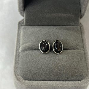 Könnte beinhalten: Ovale Ohrstecker mit einem dunklen, möglicherweise rauchigen Edelstein, der in silberfarbenem Metall gefasst ist. Die Ohrringe werden in einer grauen Samt-Schmuckschatulle präsentiert, ideal zum Verschenken.