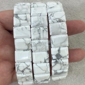 Peut inclure: Un bracelet en howlite blanc et gris. Le bracelet est composé de perles rectangulaires avec un motif marbré. Les perles sont enfilées pour former un bracelet souple. Le bracelet est présenté sur un fond neutre.