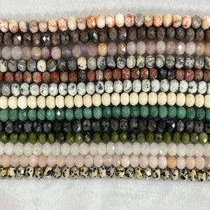 Könnte beinhalten: Eine Sammlung facettierter Edelsteinperlen in verschiedenen Farben, darunter Weiß, Rosa, Grün und Schwarz. Die Perlen sind in parallelen Reihen aufgereiht und zeigen eine Vielzahl von Texturen und Farbtönen. Ideal für die Schmuckherstellung.