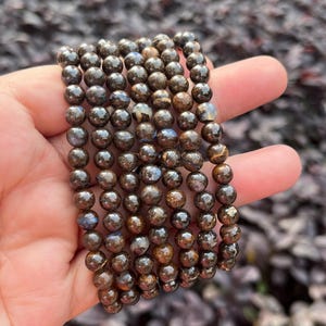 May include: A collection of beaded bracelets featuring dark brown, spherical beads with a metallic sheen. The beads are strung together, creating a layered effect. The bracelets are held in a hand, showcasing their color and texture.