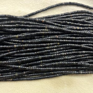 May include: Close-up of a collection of dark blue, square-shaped beads strung together. The beads have a slightly iridescent quality, with hints of lighter tones. The strands are arranged in a layered pattern, creating a textured appearance. These beads are suitable for jewelry making.