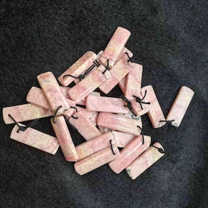 Può includere: Una collezione di pendenti rettangolari in pietra rosa con cordini neri. I pendenti hanno un aspetto marmorizzato con variazioni di colore. I cordini sono annodati nella parte superiore di ogni pendente. Sono disposti su una superficie grigio scuro.