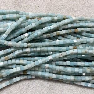 Puede incluir: Una hebra de cuentas cuadradas de color azul claro hechas de amazonita. Las cuentas tienen un acabado ligeramente iridiscente.