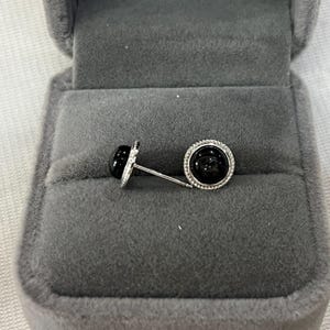 Könnte beinhalten: Ein Paar silberne Manschettenknöpfe mit einem schwarzen, kreisförmigen Stein in der Mitte. Jeder Manschettenknopf ist in einem silbernen Rahmen mit einem Ring aus kleinen, klaren Steinen gefasst. Präsentiert in einer grauen Samt-Schmuckschatulle.