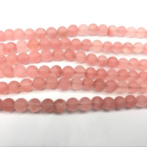 Puede incluir: Hebras de cuentas redondas de color rosa. Las cuentas tienen un acabado mate y están ensartadas, creando una línea continua. El color varía ligeramente, con algunas cuentas que aparecen más claras que otras. El fondo es blanco.