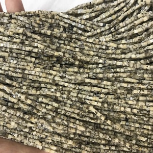 K&ouml;nnte beinhalten: Eine Str&auml;hne quadratischer Perlen aus Naturstein. Die Perlen sind hellbeige mit braunen und grauen Markierungen.