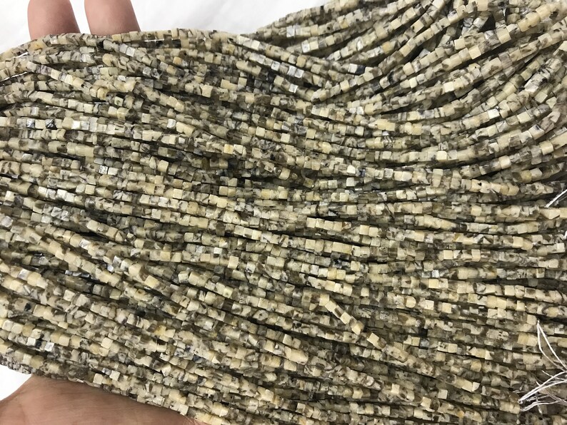 K&ouml;nnte beinhalten: Eine Str&auml;hne quadratischer Perlen aus Naturstein. Die Perlen sind hellbeige mit braunen und grauen Markierungen.