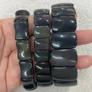 May include: Three dark grey obsidian bracelets. The bracelets are made of polished, rectangular beads. The beads have a glossy finish and are strung together with elastic. The bracelets are displayed on a light-coloured surface.
