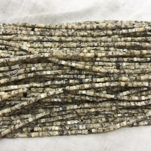 K&ouml;nnte beinhalten: Eine Str&auml;hne quadratischer Perlen aus hellem beigefarbenem Stein mit schwarzen und silbernen Flecken. Die Perlen sind etwa 4 mm gro&szlig;.