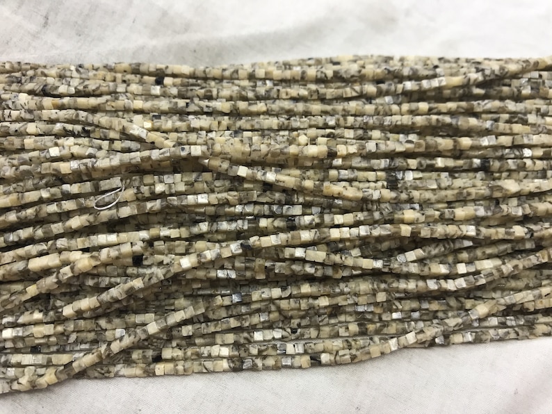 K&ouml;nnte beinhalten: Eine Str&auml;hne quadratischer Perlen aus hellem beigefarbenem Stein mit schwarzen und silbernen Flecken. Die Perlen sind etwa 4 mm gro&szlig;.