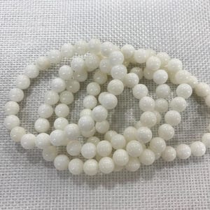 May include: Several strands of white beaded bracelets are arranged on a textured, off-white surface. The beads are round and uniform in size, creating a simple, elegant design. The bracelets are likely for jewelry.