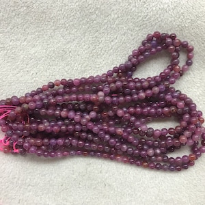 May include: A strand of purple gemstone beads, likely amethyst or ruby, with a smooth, polished finish. The beads are strung on a pink thread and are ready for jewelry making.