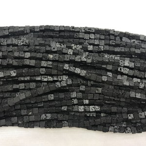 Puede incluir: Una hebra de cuentas de roca de lava negra, cada cuenta es de forma cuadrada con una superficie rugosa y texturizada.