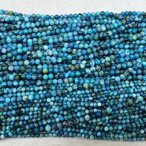 May include: A close-up of a collection of small, round beads in shades of blue and green. The beads are tightly packed, creating a textured surface. These beads are likely used for jewellery making or other crafts.