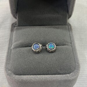 May include: A pair of silver stud earrings with a blue opal gemstone in each. The earrings are in a gray velvet jewelry box.