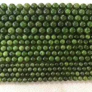 Puede incluir: Primer plano de hilos de cuentas de jade verde. Las cuentas son de diferentes tamaños y tienen un acabado pulido y brillante. El color varía de verde claro a oscuro, con algunas cuentas que muestran manchas más oscuras.