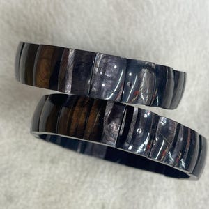 Op de afbeelding: Twee donkere houten armbanden met een glanzende afwerking. De armbanden zijn gemaakt van rechthoekige segmenten, met zichtbare houtnerf in tinten bruin en zwart. De armbanden zijn op elkaar gestapeld.