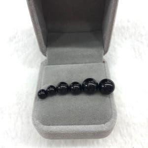 Puede incluir: Un conjunto de seis esferas negras brillantes de diferentes tamaños, dispuestas en fila dentro de una caja de joyería de terciopelo gris. Las esferas varían de aproximadamente 0,6 cm a 1,9 cm de diámetro, mostrando una gradación de tamaños.