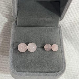 May include: A pair of rose quartz earrings in a gray velvet jewelry box. The earrings are round and have a smooth, polished finish.