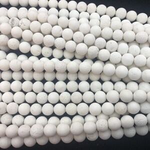 May include: A close-up shot of numerous white, spherical beads with a textured surface. The beads are tightly packed together, filling the image. They appear to be made from a porous material, and are likely used for jewellery making.