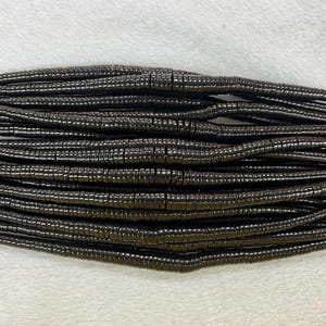 May include: A strand of black hematite beads, each bead is a small cylinder with a slightly rounded edge. The beads are arranged in a neat, parallel pattern.