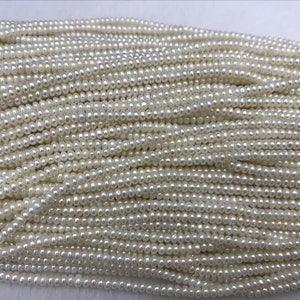 Peut inclure: Gros plan sur de nombreux brins de petites perles blanches crémeuses. Les perles sont de taille et de forme uniformes, créant une surface lisse et brillante. Les brins sont disposés les uns près des autres, suggérant un approvisionnement en vrac de composants de bijoux.