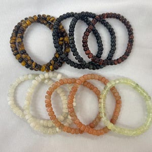 Könnte beinhalten: Eine Sammlung von zehn elastischen Perlenarmbändern, die in zwei Reihen angeordnet sind. Die obere Reihe besteht aus Tigerauge-, schwarzen und dunkelrotbraunen Perlen. Die untere Reihe besteht aus weißen, hellorangen und blassgrünen, durchscheinenden Steinperlen.