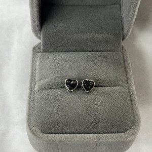 Könnte beinhalten: Ein Paar herzförmige Ohrstecker mit schwarzem Mittelteil und silberfarbenem Rand, präsentiert in einer offenen, grauen Samt-Schmuckschatulle. Die Ohrringe sind klein und zart.