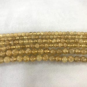 Könnte beinhalten: Nahaufnahme von mehreren Strängen goldgelber Edelsteinperlen. Die Perlen sind unterschiedlich groß, wobei die größten unten und die kleinsten oben sind. Die Perlen haben ein durchscheinendes Aussehen und eine leicht strukturierte Oberfläche. Der Hintergrund ist schlicht weiß.