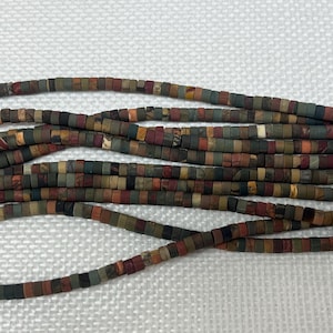 May include: Strands of multi-colored, flat, rectangular beads in shades of brown, green, red, and gray. The beads are strung together, creating a textured and earthy appearance. Ideal for jewelry making.